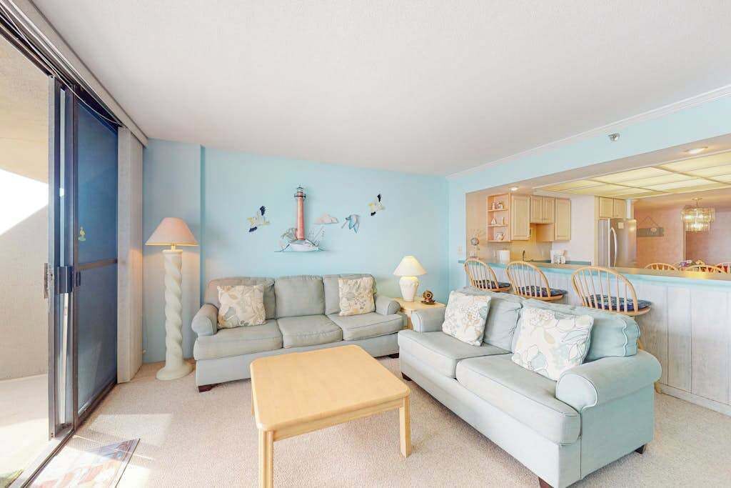A living room with a sofa, a coffee table, and a kitchen area visible through an open door.