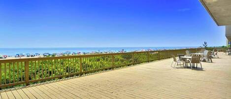A wooden deck with outdoor seating, overlooking a beach with umbrellas and a clear sky.