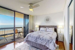 A bedroom with a bed, a ceiling fan, a nightstand, a lamp, a decorative wall piece, and a view of the beach through a sliding door. - Wonderful Suite in Beachfront Complex w/Shared Pool, Free WiFi, Private W/D (Ocean City)