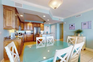 A modern kitchen with wooden cabinets, a stainless steel refrigerator, and a glass dining table with white chairs. - Belmont Towers 708 (Ocean City)