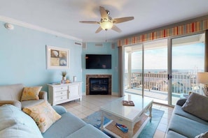A living room with a TV, fireplace, ceiling fan, and large windows with a city view. - Belmont Towers 708 (Ocean City)