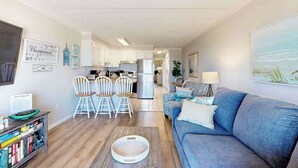 Ein modernes Wohnzimmer mit einer Couch, einem Couchtisch und einem Bücherregal. Es gibt einen Essbereich mit Stühlen und einem Tisch. Im Hintergrund ist eine Küche erkennbar.