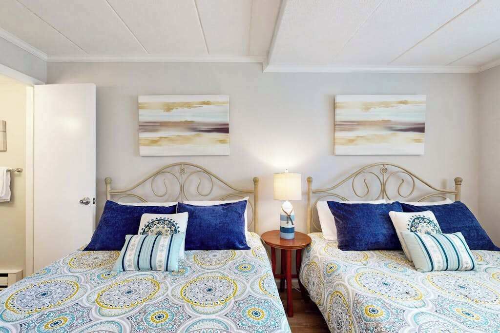 A room with two beds, each with patterned bedding, a bedside table with a lamp, and abstract wall art.