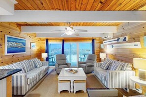 A room with wooden walls, a ceiling fan, two sofas, a coffee table, and a view through large windows. - Beachwalk 6 (Ocean City)