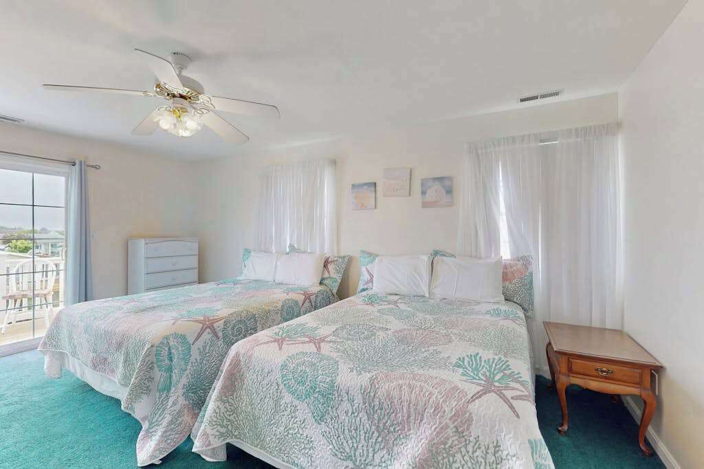 A bedroom with two beds, a ceiling fan, a window with curtains, a small wooden nightstand, and a balcony with a view.