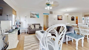 Un salon comprenant un canapé, une table à manger avec des chaises, une table basse blanche, une chaise blanche avec un coussin, un ventilateur de plafond et une bibliothèque.