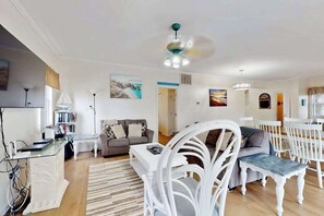 Ein Wohnzimmer mit einer Couch, einem Esstisch mit Stühlen, einem weißen Couchtisch, einem weißen Sessel mit Kissen, einem Deckenventilator und einem Bücherregal.