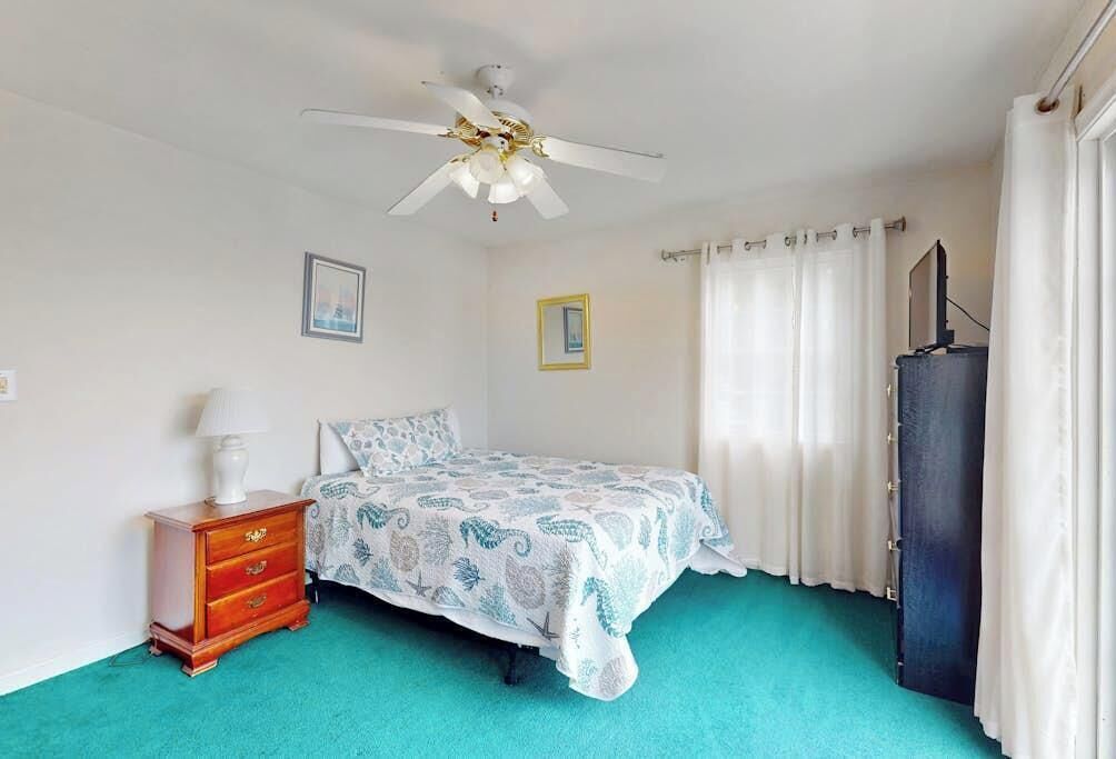 Un dormitorio con cama, mesita de noche, ventilador de techo, televisión y cortinas.