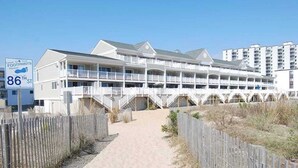 Un hotel frente a la playa con varios balcones, un paseo marítimo de arena y una cerca de madera.