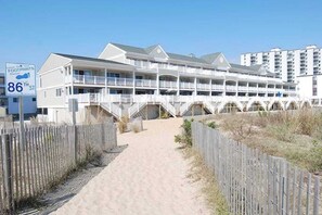 Ein Strandhotel mit mehreren Balkonen, einem Sandweg und einem Holzzaun.