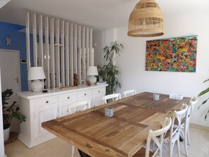 Ein Essbereich mit Holztisch, weißen Stühlen, einem weißen Schrank mit Schubladen, einer hängenden Korbellampe und einem farbenfrohen, abstrakten Gemälde an der Wand.