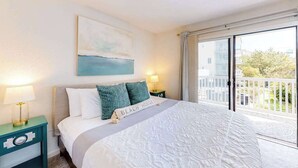 A bedroom with a bed, a nightstand, a lamp, a painting, a balcony with a railing, and a view of buildings and greenery.