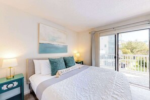 A bedroom with a bed, a nightstand, a lamp, a painting, a balcony with a railing, and a view of buildings and greenery. - Sea Mark 208 (Ocean City)