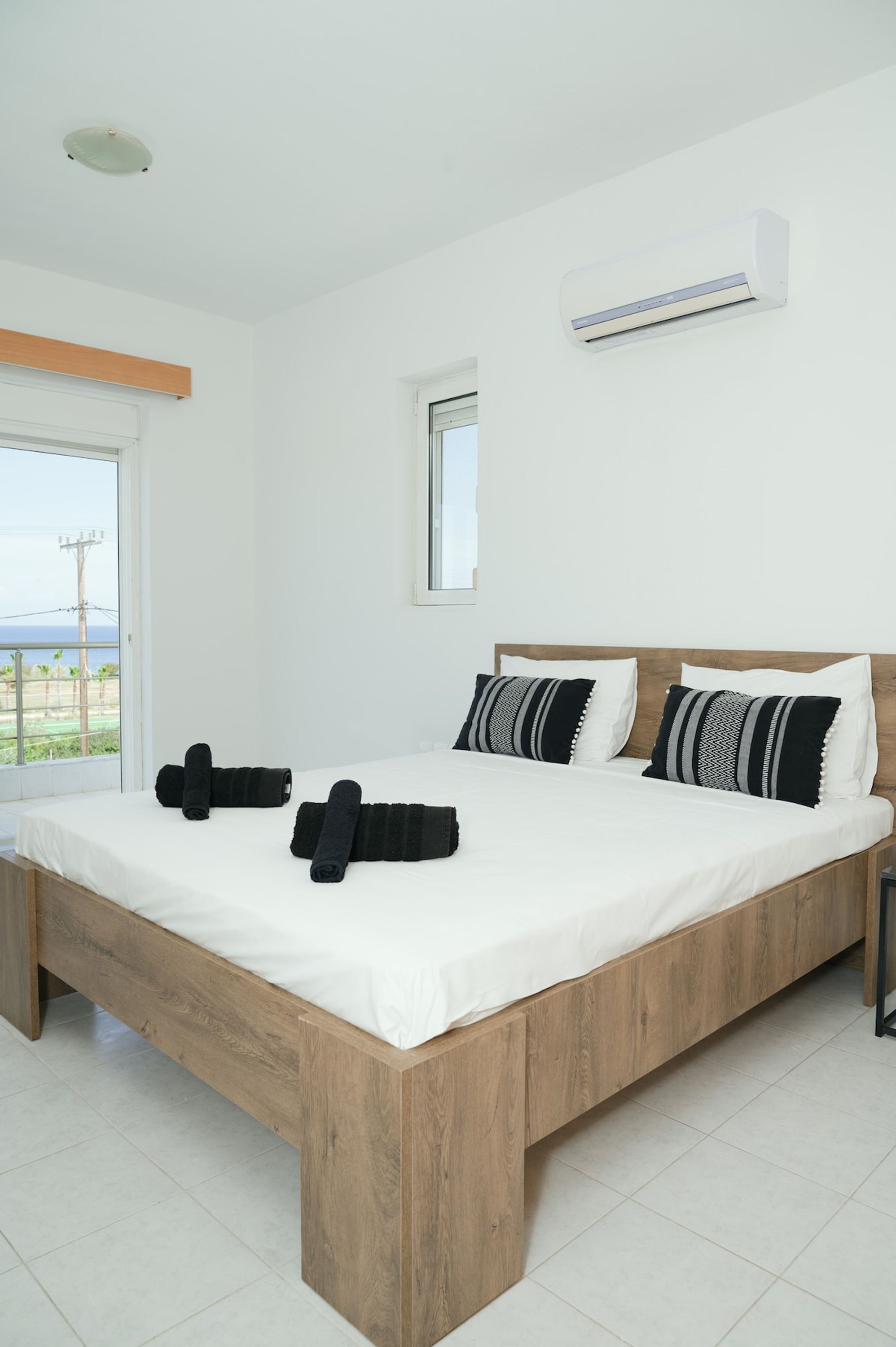 Une chambre avec un lit en bois, du linge de lit blanc, des serviettes noires et un appareil de climatisation.