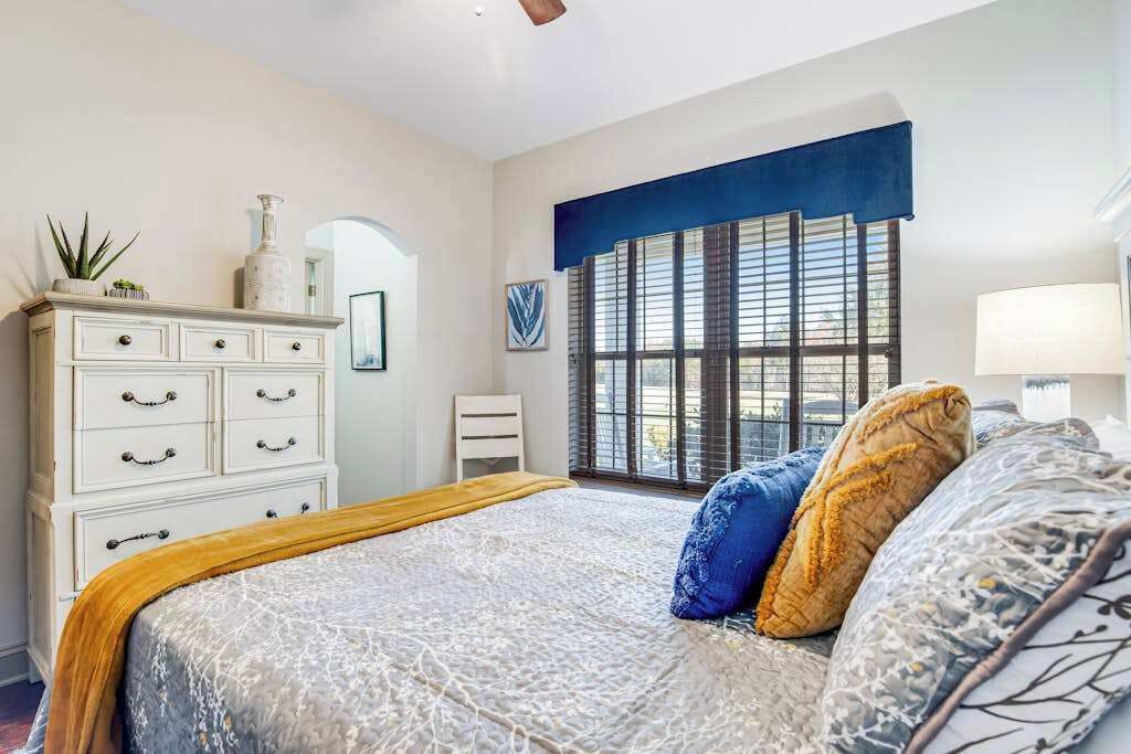 Une chambre à coucher avec un lit, une commode, une fenêtre avec des stores et un ventilateur de plafond.