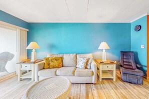 A living room with a blue wall, a sofa, two nightstands with lamps, a recliner, and a wicker chair. - Oceanfront Condo with Beach Views, WiFi, Washer-Dryer, Parking & Walkable Locale (Ocean City)