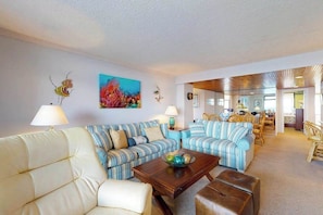 A living room with striped sofas, a coffee table, and a dining area with wicker furniture. - Cozy condo in beachfront complex with pool & W/D (Ocean City)