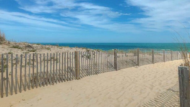Ein sandiger Strand mit einer Holzlattenveranda und blauem, wolkenlosem Himmel.