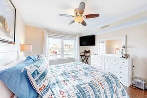 A hotel room with a bed, a ceiling fan, a television, a dresser, and a window with a view of buildings. - Bayspot L-101 (Ocean City)