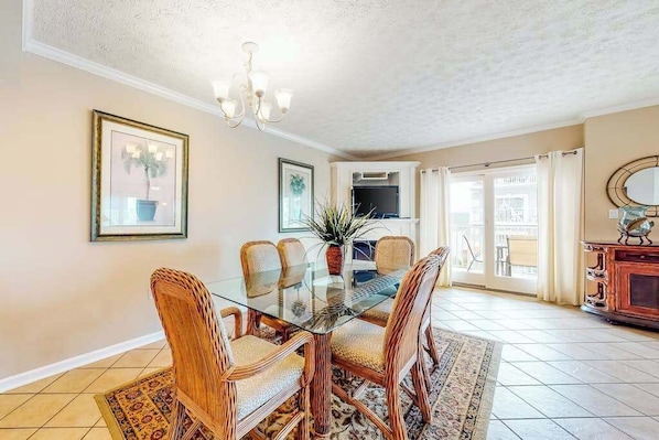 A dining area with a glass table, wicker chairs, a patterned rug, a chandelier, and a view to a balcony. - Bay & ocean views at Sea Palms - free WiFi, private washer/dryer & dog-friendly (Ocean City)