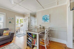 A living room with a sofa, a dining table with chairs, a bookshelf, and a door leading to another room. - Walk Everywhere - Cute Coastal Cottage - Alfresco Dining, Outdoor Shower & Porch (Ocean City)