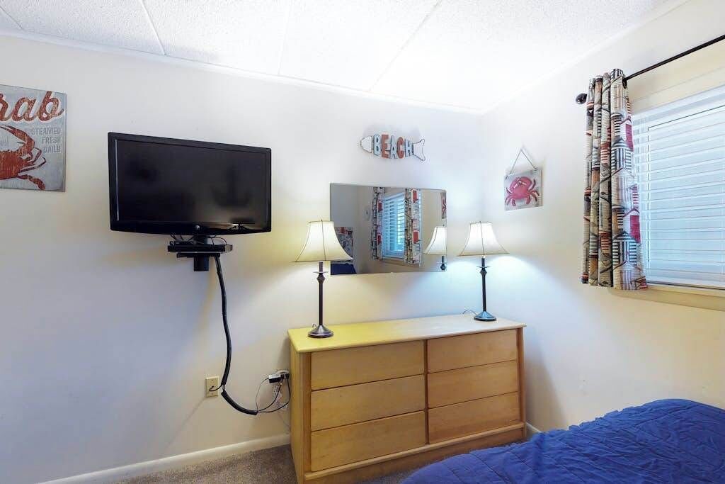 Een slaapkamer met een aan de muur bevestigde televisie, een dressoir met twee lampen, een spiegel en een bed met een blauwe deken.