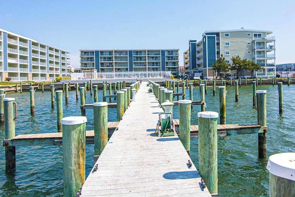 A wooden pier leading to a waterfront hotel with multiple balconies and a pool area.