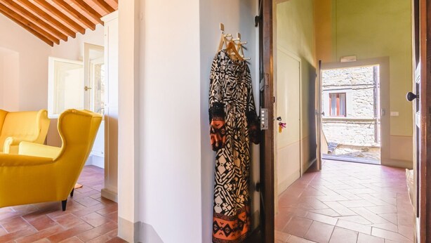 A hallway with a yellow chair, a patterned garment hanging on a rack, and a door leading to an outdoor area. - Il Ponte - a romantic retreat blending history and panoramic views (Cortona)
