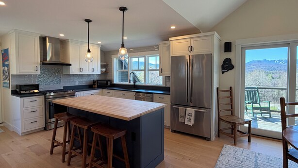 A modern kitchen with a central island, bar stools, stainless steel appliances, pendant lighting, and a view through large windows. - Cozy 1-bedroom apartment in Stowe with amazing views, AC & gas fireplace (Stowe)