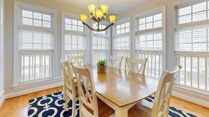 A dining room with a wooden table, white chairs, a chandelier, and large windows with white shutters. - Beach Retreat A (Ocean City)