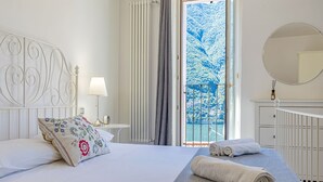 A hotel room with a bed, a bedside table with a lamp, a window with a view of snow-covered trees, and a white dresser. - Angela Lake View Apartment (Pognana Lario)