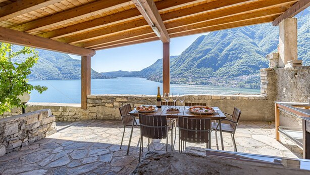 A covered outdoor dining area with stone flooring, wooden beams, and a table set for two with plates of food and wine glasses. - Angela Lake View Apartment (Pognana Lario)