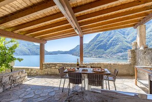A covered outdoor dining area with stone flooring, wooden beams, and a table set for two with plates of food and wine glasses.