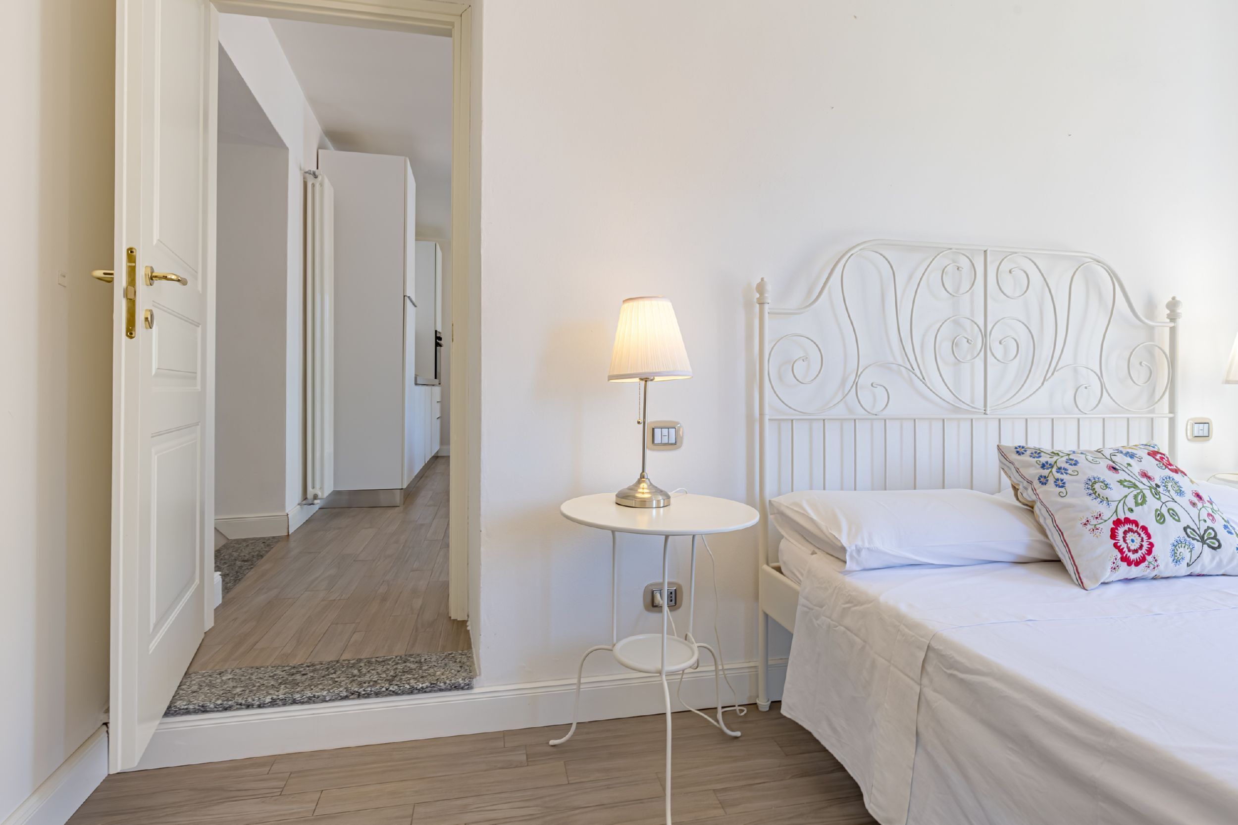 Un dormitorio con una cama, una mesita de noche con lámpara y una puerta que da a otra habitación.