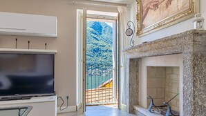 A room with a TV, a fireplace, a balcony with a view of a mountain, and a painting on the wall. - Angela Lake View Apartment (Pognana Lario)