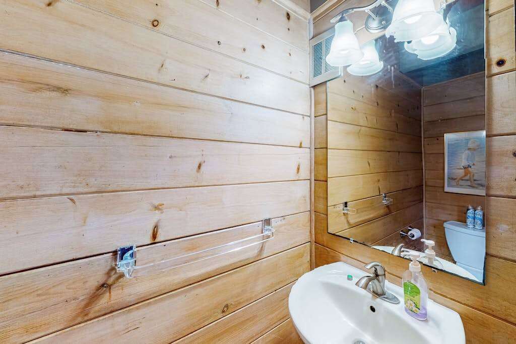Une salle de bain aux murs lambrissés, avec un lavabo blanc, un miroir et des luminaires au plafond.