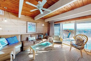 Un salon chaleureux avec un plafond en bois, un ventilateur de plafond, une table basse en verre, un canapé en cuir marron et une grande fenêtre offrant une belle vue.