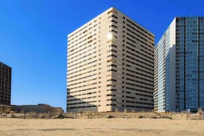 A high-rise building with a grid of windows, a flat roof, and a sandy ground in front.