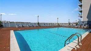 A swimming pool with a wooden deck, lounge chairs, and a building with balconies. - Assateague House 511 (Ocean City)