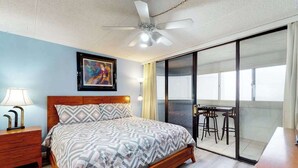 A bedroom with a bed, a nightstand, a ceiling fan, a painting, a sliding glass door leading to a balcony with a table and chairs. - Well-Appointed Vacation Getaway in Oceanfront Complex w/Shared Pool, Free WiFi (Ocean City)