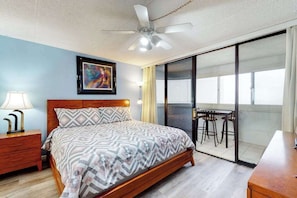 A bedroom with a bed, a nightstand, a ceiling fan, a painting, a sliding glass door leading to a balcony with a table and chairs. - Well-Appointed Vacation Getaway in Oceanfront Complex w/Shared Pool, Free WiFi (Ocean City)