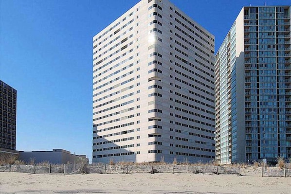 A tall, modern building with a grid of windows, surrounded by a sandy area with sparse vegetation. - Well-Appointed Vacation Getaway in Oceanfront Complex w/Shared Pool, Free WiFi (Ocean City)