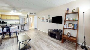 A modern living room with a flat-screen TV, a wooden bookshelf, and a dining area with a table and chairs. - Well-Appointed Vacation Getaway in Oceanfront Complex w/Shared Pool, Free WiFi (Ocean City)