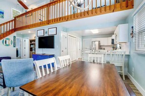 A dining area with a wooden table and white chairs, a kitchen with white cabinets, and a staircase with wooden railings.