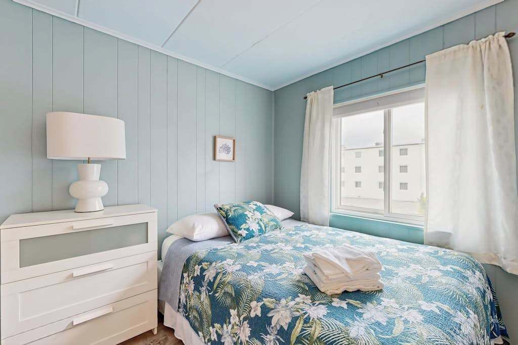 A bedroom with a bed, a nightstand, a lamp, a window with curtains, and a view of buildings outside.