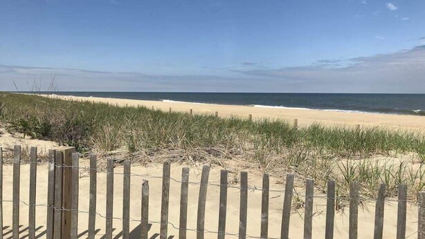 A sandy beach with a wooden fence and grass in the foreground. - 3rd-floor condo near the beach with balcony, partial ocean views, & central AC (Ocean City)
