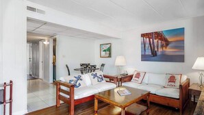 A room with a wooden floor, white walls, a white sofa with anchor pillows, a wooden coffee table, a painting of a pier, and a lamp. - 3rd-floor condo near the beach with balcony, partial ocean views, & central AC (Ocean City)