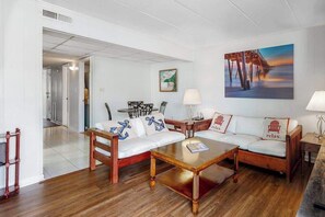 A room with a wooden floor, white walls, a white sofa with anchor pillows, a wooden coffee table, a painting of a pier, and a lamp. - 3rd-floor condo near the beach with balcony, partial ocean views, & central AC (Ocean City)