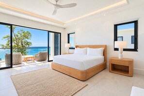 A modern bedroom with a large bed, two bedside tables, a ceiling fan, and a view of the ocean. - Beach Front Staffed 6br Villa | Lux Resort by Bocobay (St. Ann’s Bay)