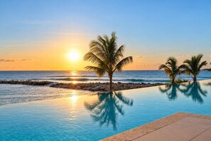 A clear view of a swimming pool with a sunset in the background, palm trees, and a rocky shoreline. - Beach Front Staffed 6br Villa | Lux Resort by Bocobay (St. Ann’s Bay)